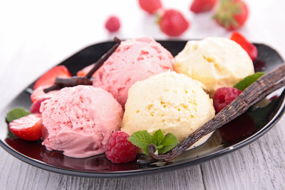 A black plate with four scoops of pink and yellow ice cream, garnished with strawberries, raspberries, mint leaves, and vanilla beans.