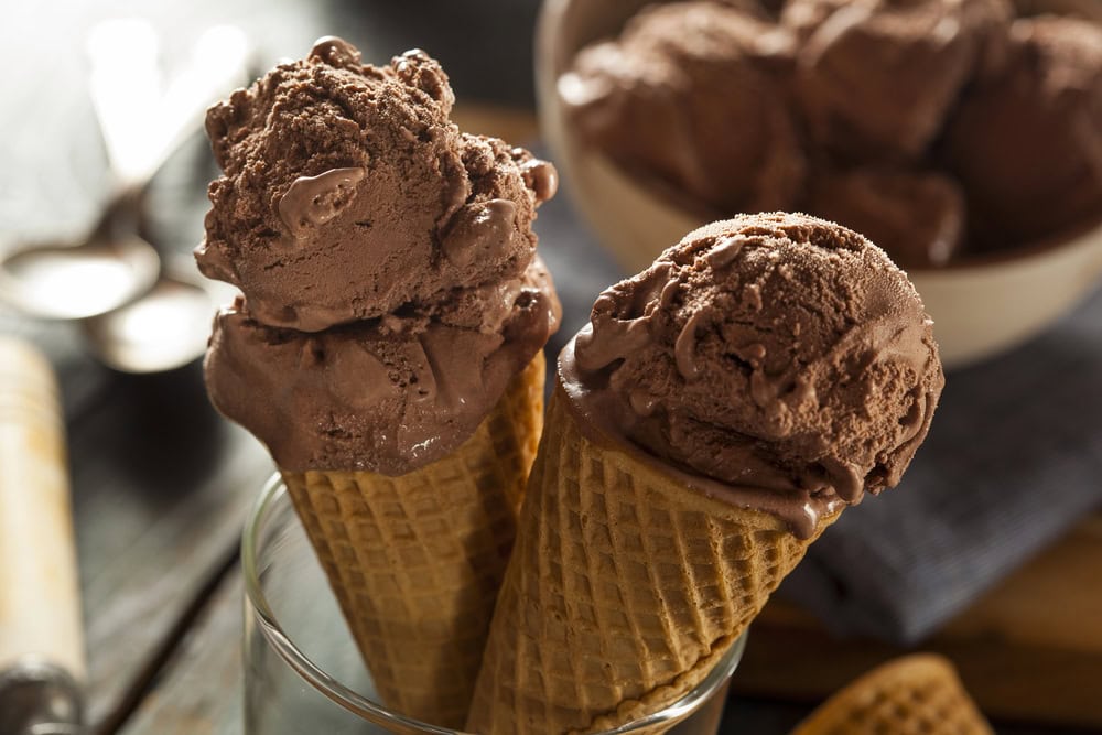 Two waffle cones filled with scoops of chocolate ice cream in a glass, with a bowl of more chocolate ice cream and spoons in the background.