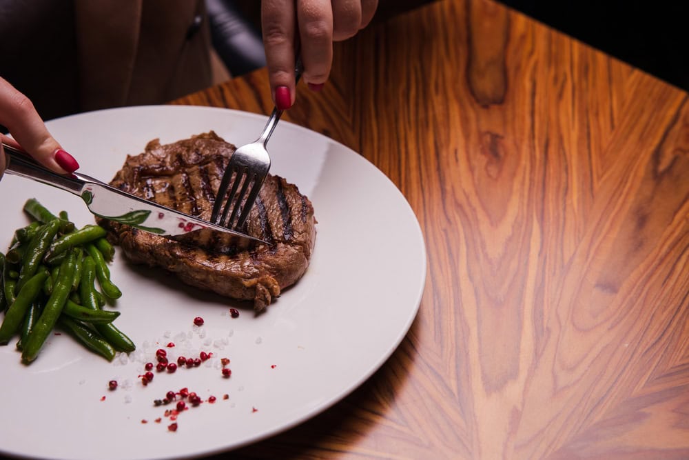 A person cuts a grilled steak on a white plate with green beans and red peppercorns, on a wooden table.
