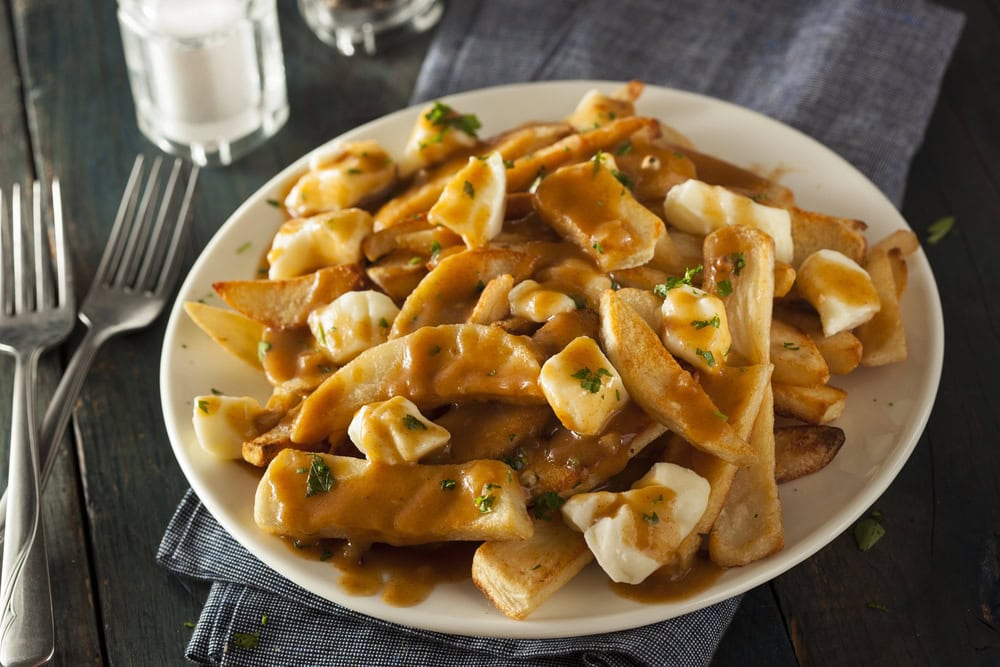 A plate of poutine with french fries, cheese curds, and brown gravy, garnished with chopped herbs, on a dark wooden table with two forks and salt and pepper shakers nearby.