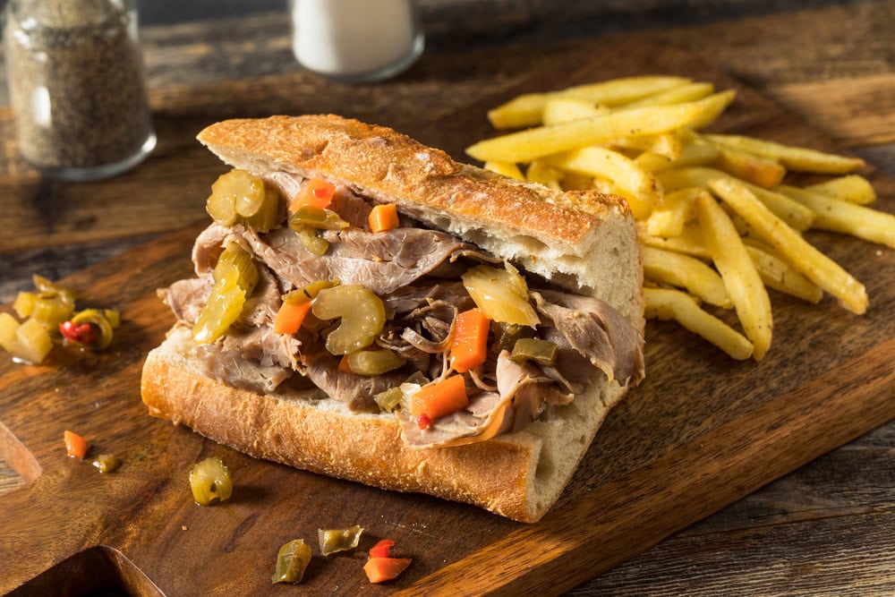 A sandwich filled with sliced beef and pickled vegetables sits on a wooden board next to a serving of French fries.