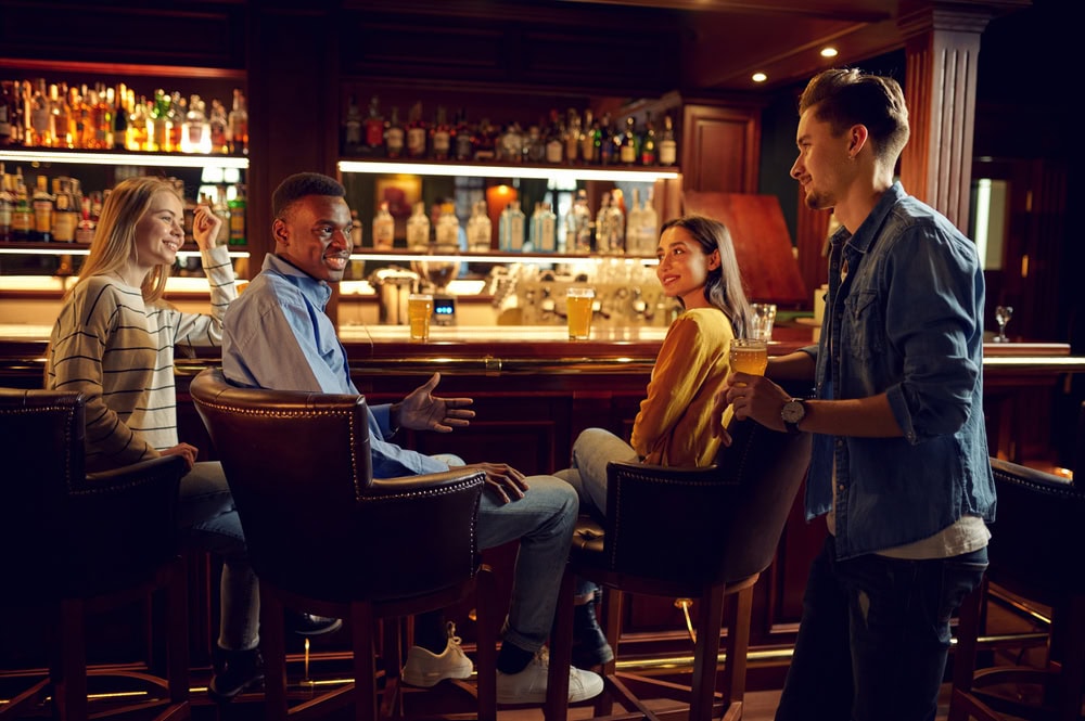 Four people sit and stand at a bar, talking and smiling, with drinks on the counter and shelves of bottles in the background.