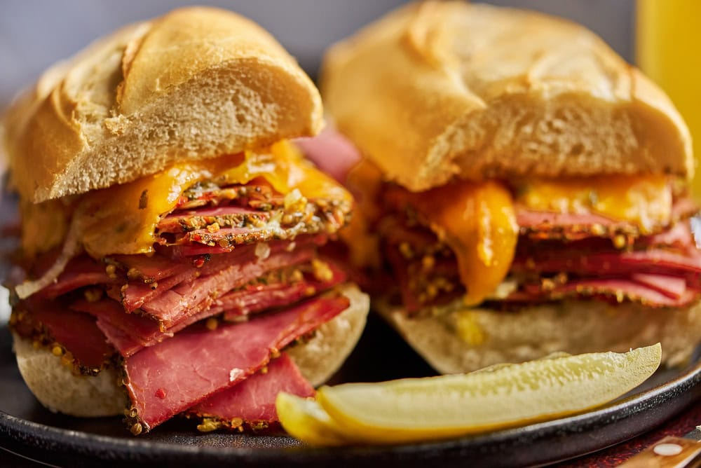 Two halves of a sandwich filled with layers of pastrami and melted cheddar cheese on a plate, with a pickle wedge in front.