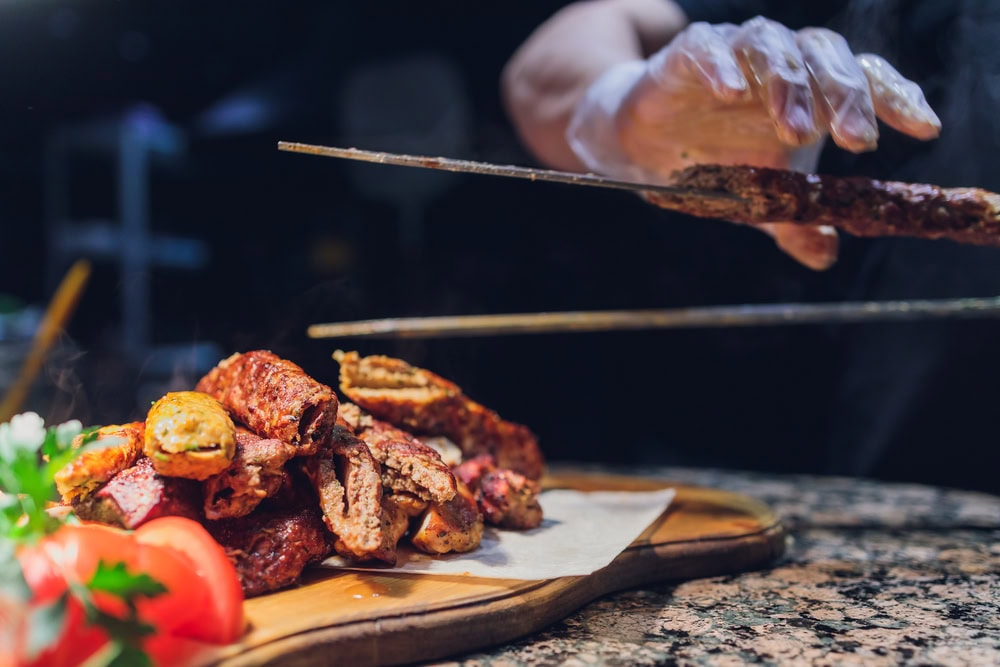 A person wearing a glove is placing grilled meat skewers onto a wooden serving board with sliced tomatoes and parsley.