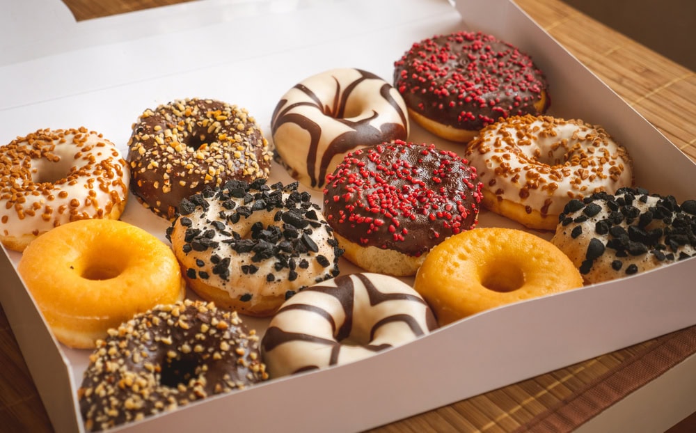 A white box filled with twelve assorted donuts, including plain, chocolate, vanilla, and donuts topped with sprinkles, nuts, and cookie crumbs, arranged in three rows.