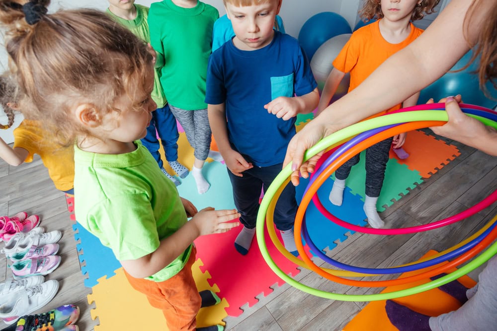 A group of children stands on colorful foam mats while an adult holds several colorful hula hoops in a classroom or play area.