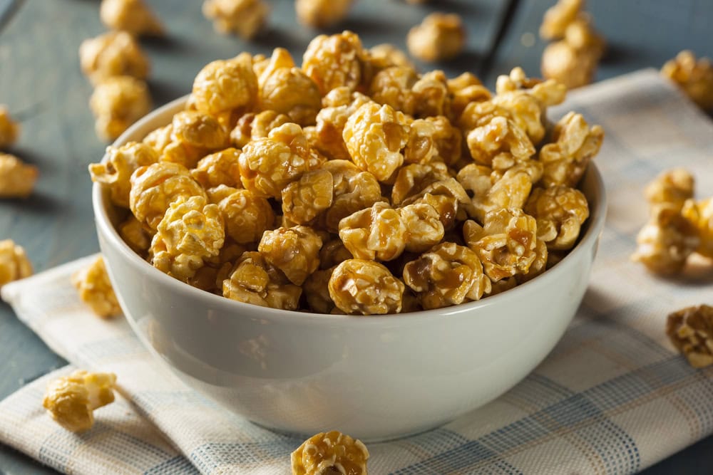 A white bowl filled with caramel-coated popcorn sits on a plaid cloth, with a few pieces scattered around on a wooden surface.
