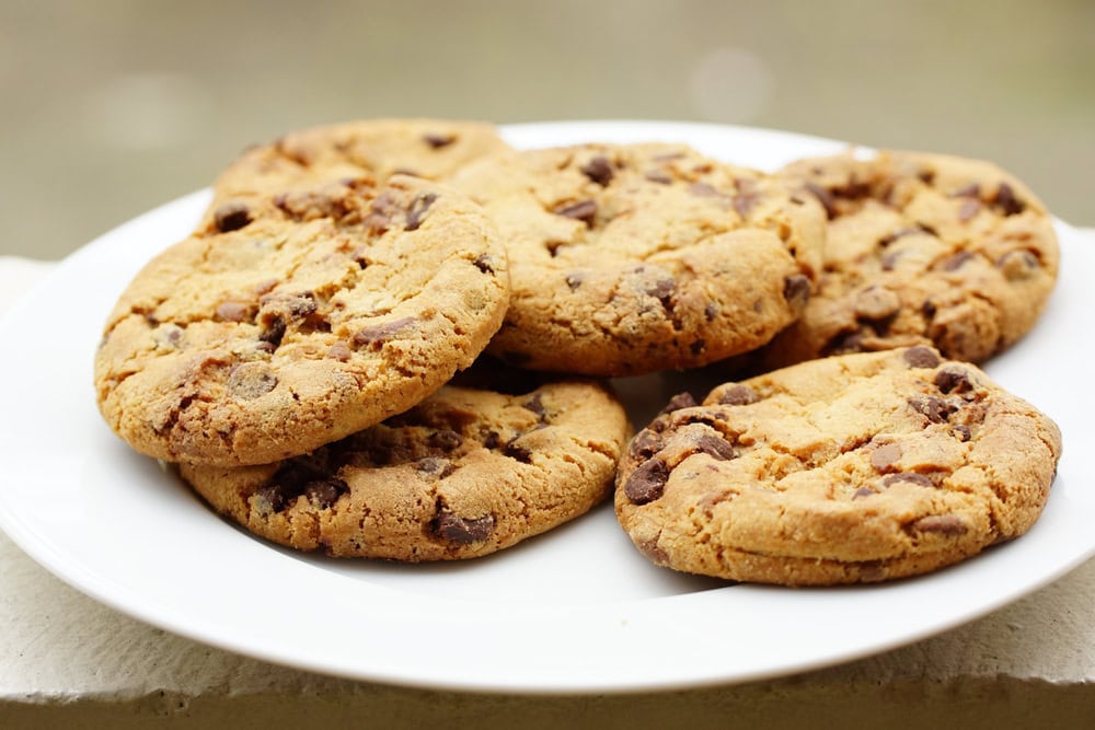 A white plate with six chocolate chip cookies stacked on it, set against a blurred background.