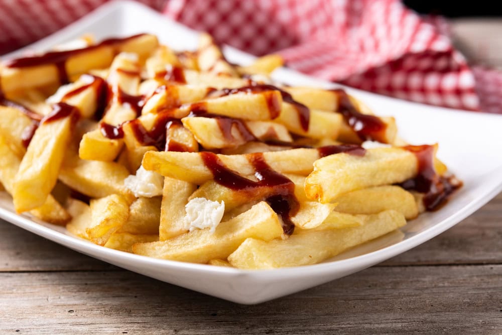 A plate of French fries topped with barbecue sauce, served on a white dish with a red checkered cloth in the background.