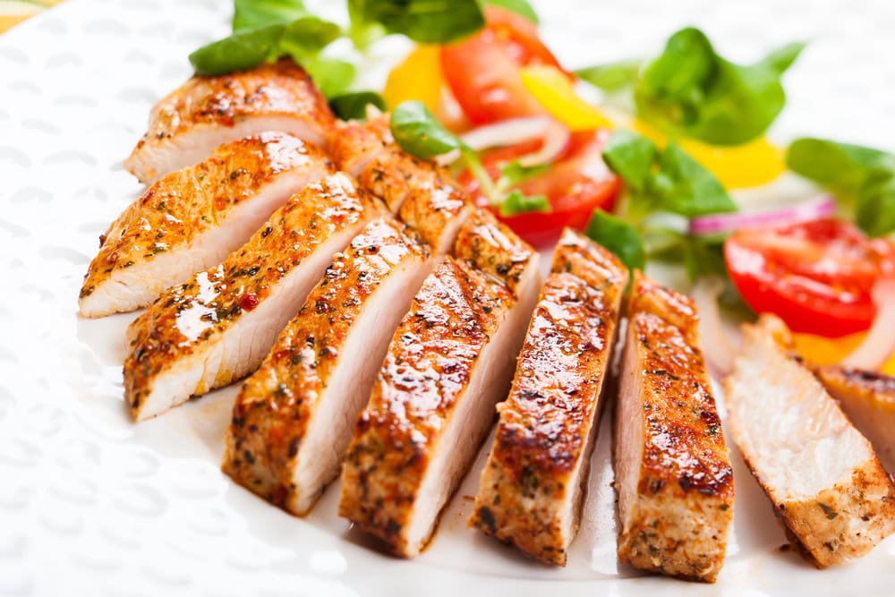 Sliced grilled chicken breast on a white plate, served with a side salad of tomatoes, greens, and yellow bell peppers.