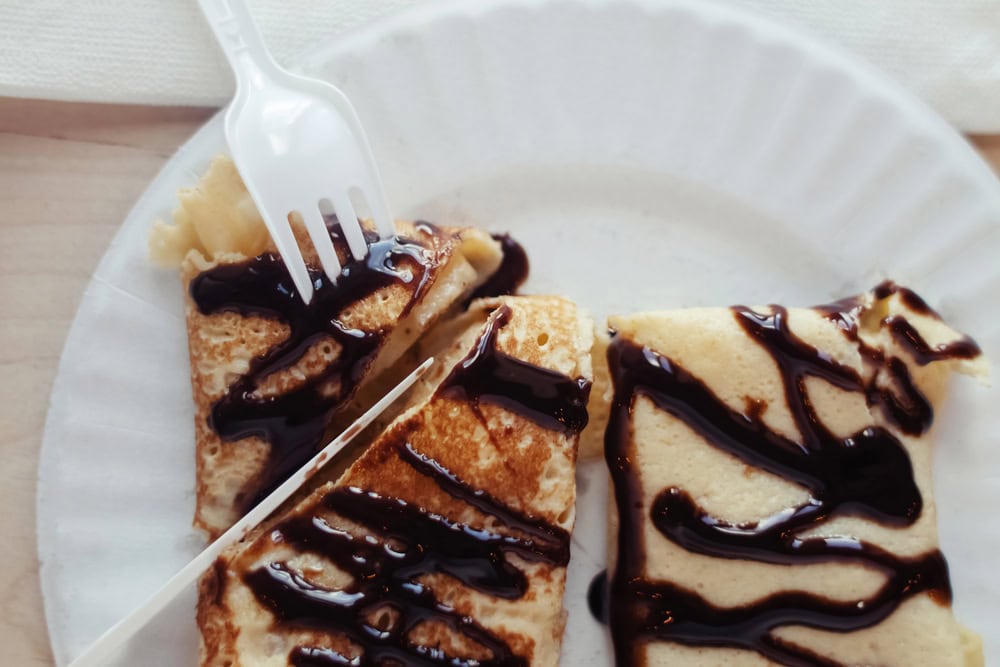 Two folded crepes with chocolate sauce on a paper plate, with a plastic fork and wooden stick placed on top.