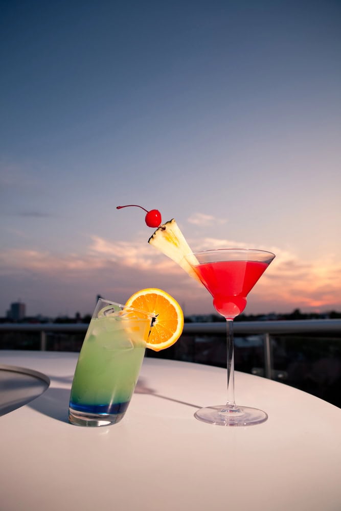 Two colorful cocktails on a white table at sunset; one is green with an orange slice, the other is pink with a cherry and pineapple garnish in a martini glass.