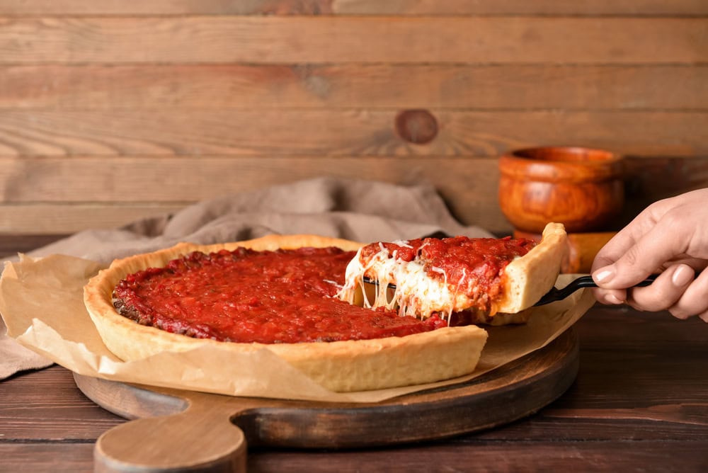 A hand lifts a slice of deep-dish pizza with tomato sauce and melted cheese from a whole pizza on a wooden serving board.