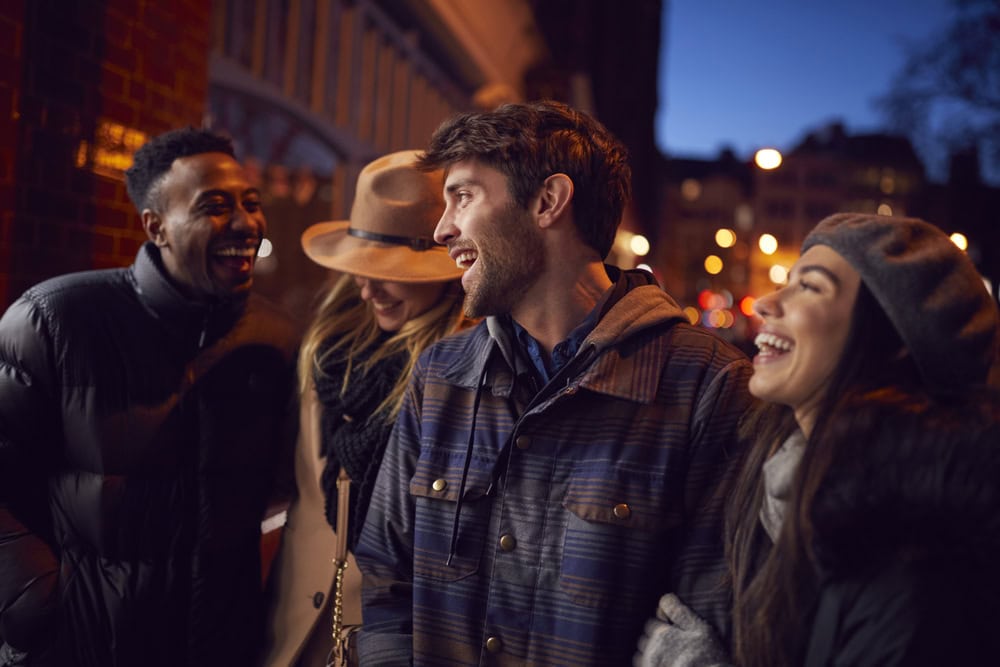 Four people dressed in winter clothing stand close together on a city street at dusk, smiling and laughing with blurred lights in the background.