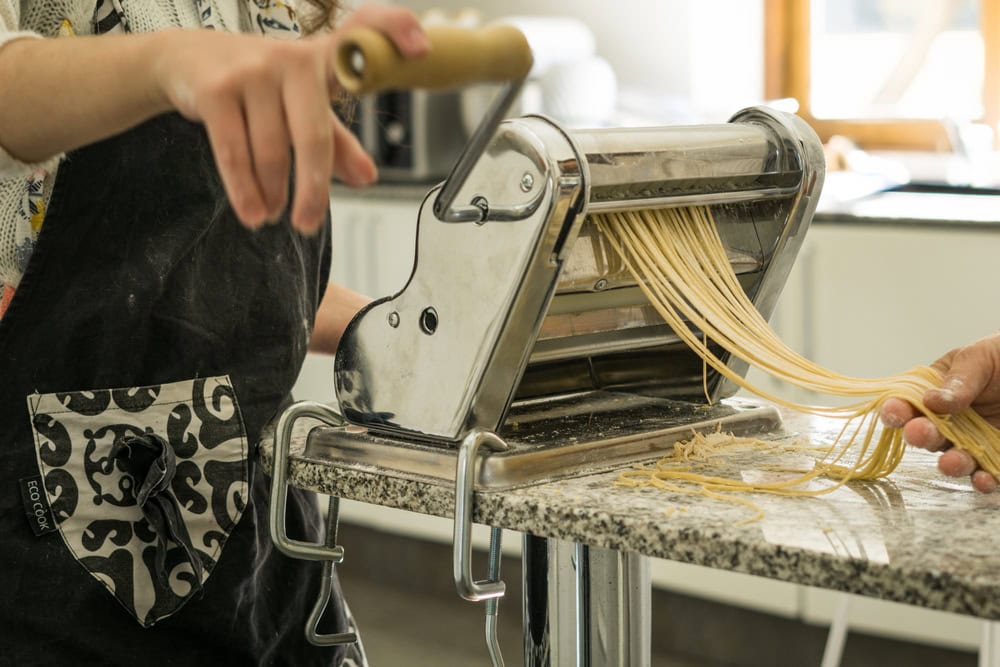 Person using a pasta machine to roll out and cut fresh pasta dough on a kitchen counter.