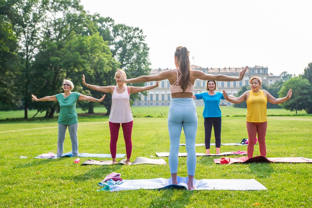 A yoga instructor leads a group of four women in an outdoor yoga class on mats in a grassy park with a building in the background.