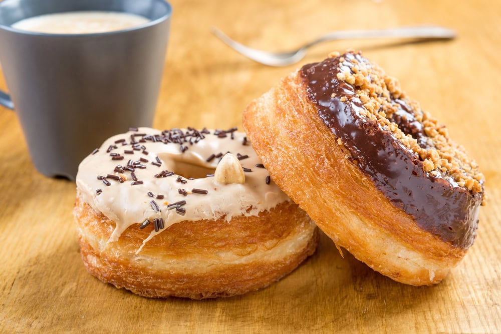 Two cronut pastries, one with white icing and chocolate sprinkles, the other with chocolate icing and nuts, sit on a wooden surface near a cup of coffee.