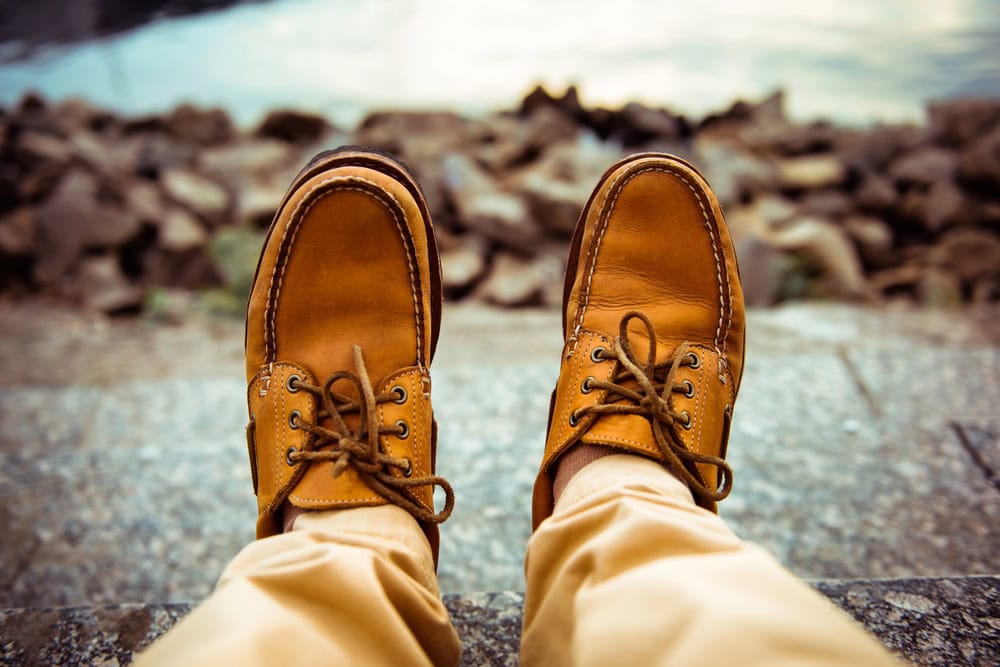 A person wearing tan leather lace-up shoes and khaki pants sits on stone steps near a rocky waterfront.