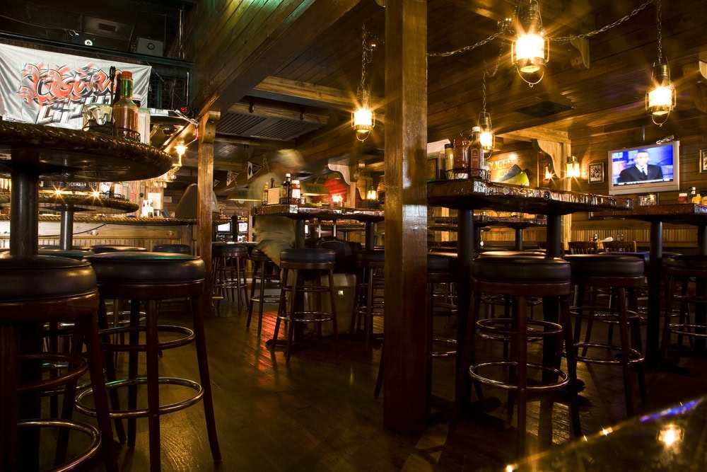 A dimly lit bar interior with high wooden tables and stools, hanging lanterns, a TV on the wall, and bottles on the tables.