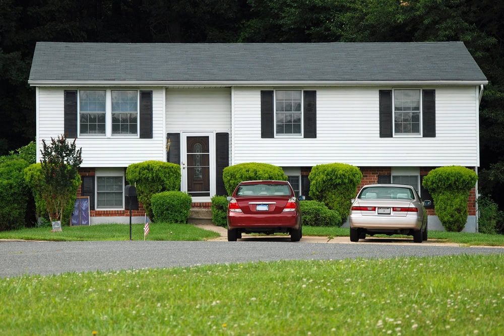 A two-story suburban house with white siding, black shutters, and a central entrance. Two cars are parked in the driveway, and trimmed bushes line the front yard.