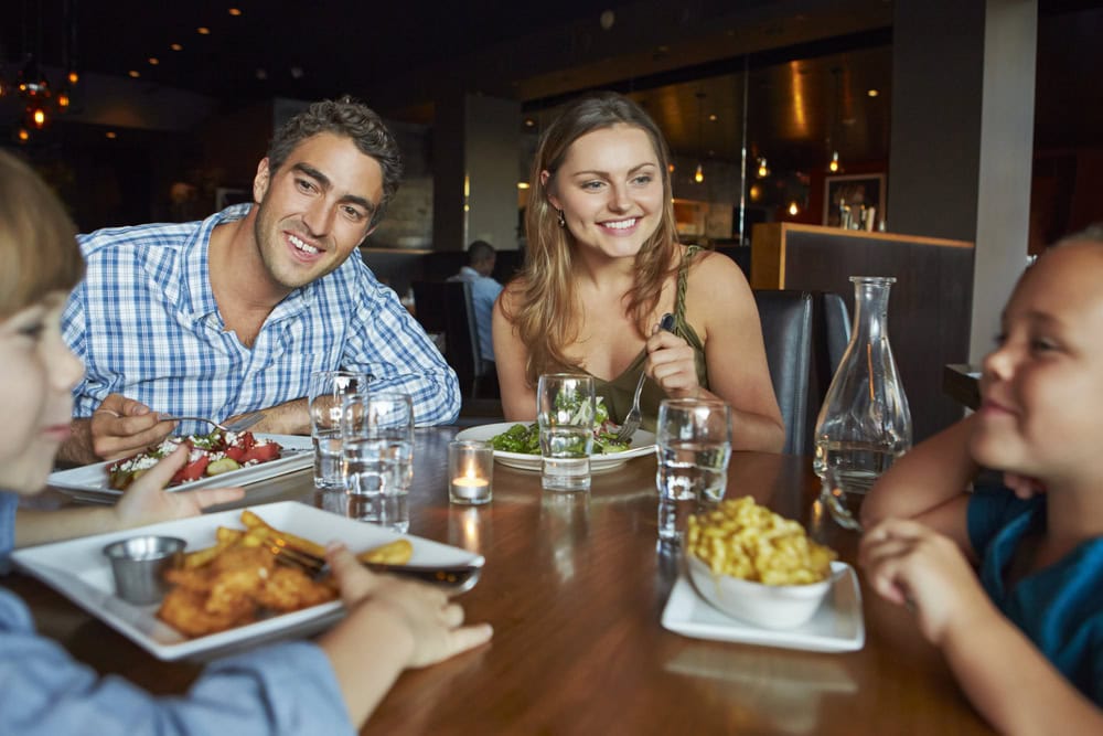 Two adults and two children are seated at a restaurant table, smiling and eating dinner together. Plates of food and glasses of water are in front of them.