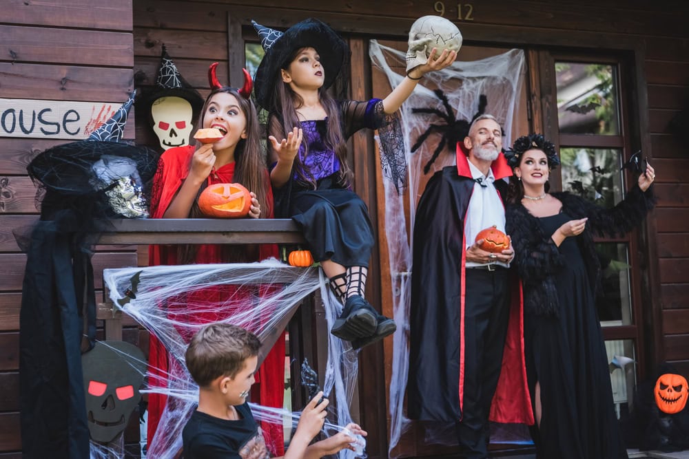 A group of people in Halloween costumes pose on a decorated porch with pumpkins, cobwebs, and skull props.