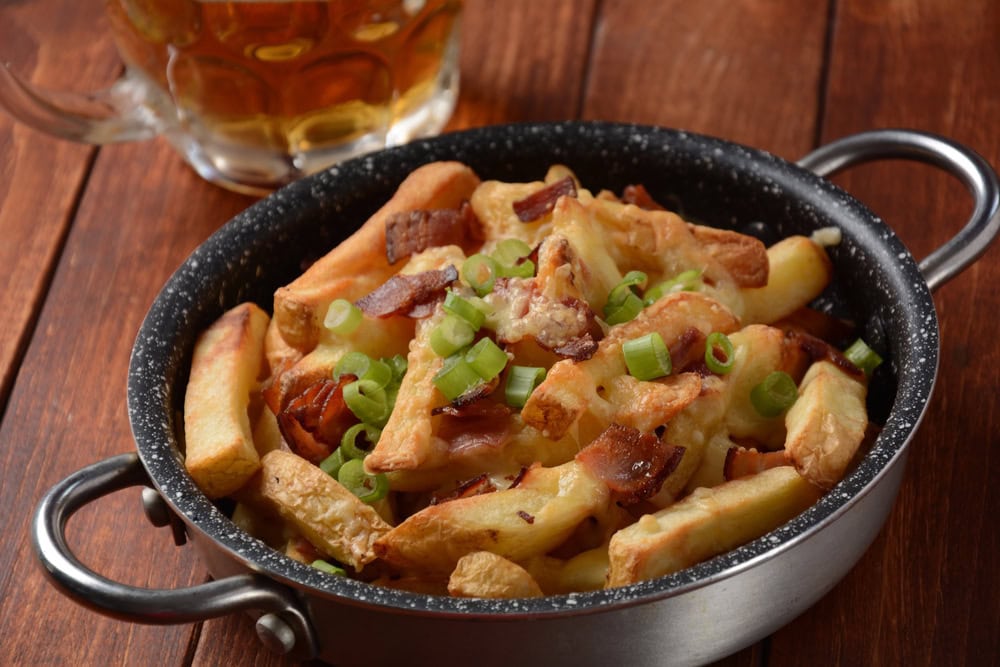 A skillet of loaded fries topped with melted cheese, chopped bacon, and sliced green onions sits on a wooden table next to a glass of beer.