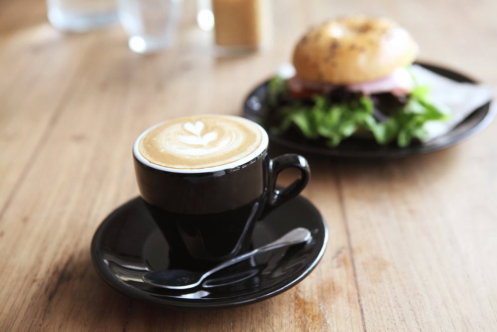 A black cup of latte with latte art sits on a saucer with a spoon, next to a plate with a bagel sandwich on a wooden table.