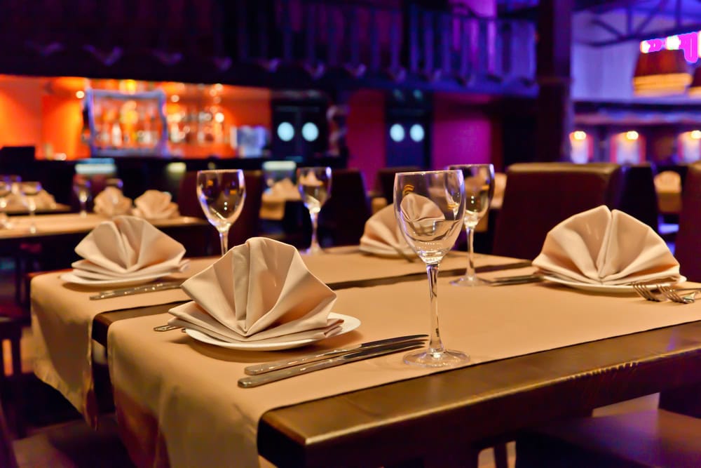 A restaurant dining table is set with folded white napkins, wine glasses, plates, and cutlery. Other similarly set tables and a bar are visible in the background.