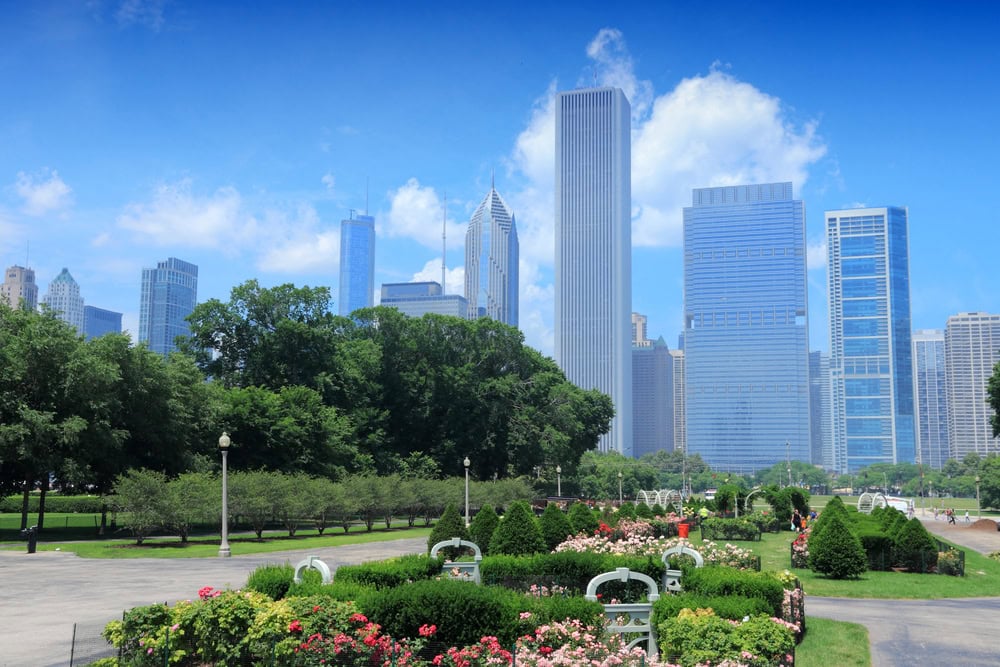Downtown Chicago skyline with modern skyscrapers, seen from a park with green lawns, trees, flower beds, and benches under a blue sky with scattered clouds.