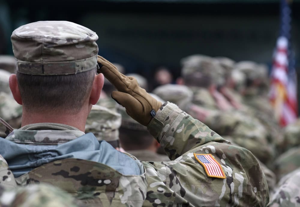A group of U.S. soldiers in camouflage uniforms, one saluting, with an American flag patch visible on a sleeve.