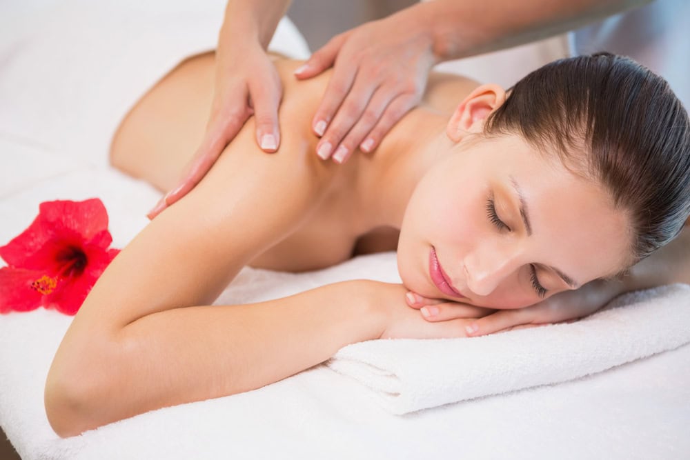 A woman receives a massage on a white massage table, with a red hibiscus flower nearby.