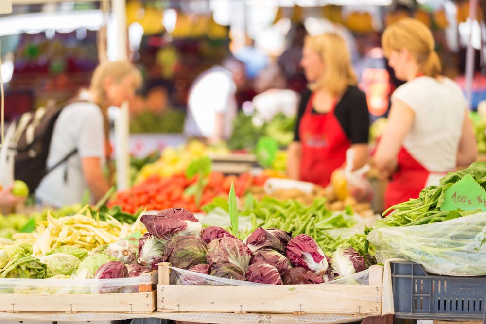 Fresh vegetables displayed on a market stall with three people, two wearing red aprons, interacting in the background.