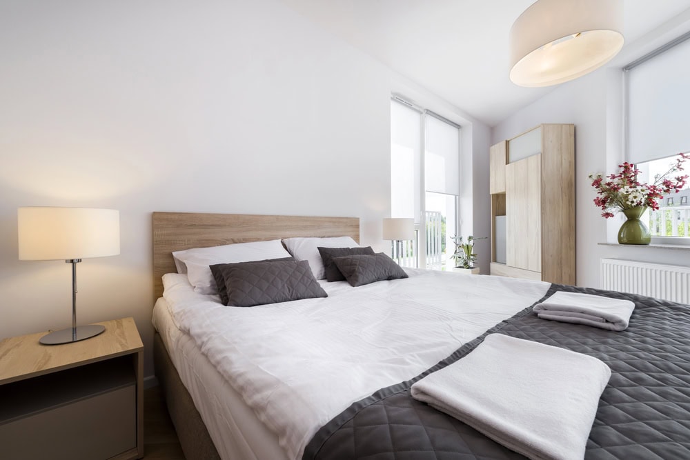 Modern bedroom with a large bed, two bedside tables with lamps, a wardrobe, and a window with blinds. The bed is neatly made with gray pillows and folded towels.