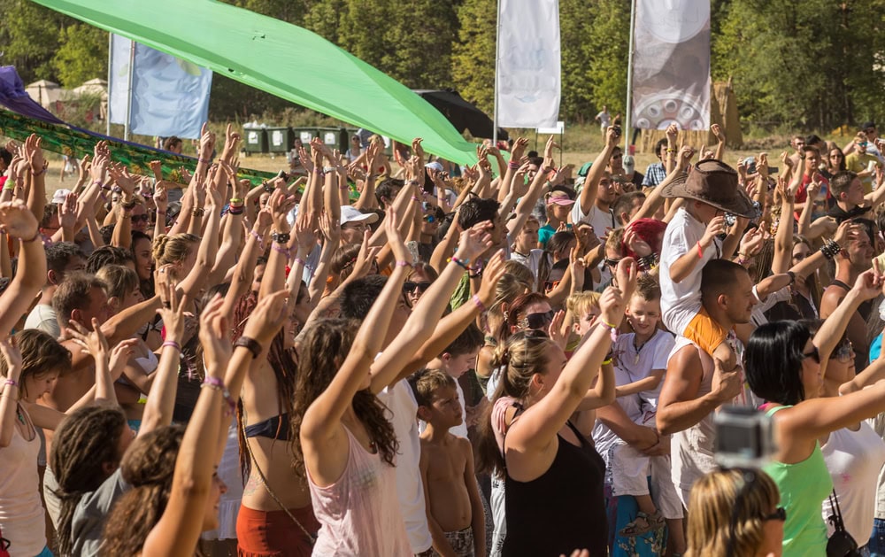 A large crowd of people outdoors with raised arms, some dancing and others standing under green shade cloths, at a daytime festival or event.