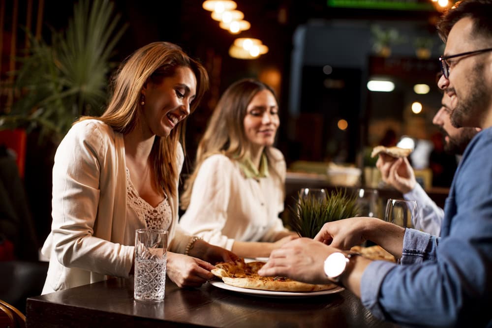 Three people sit at a table in a restaurant, sharing a pizza and drinks, smiling and engaging in conversation.