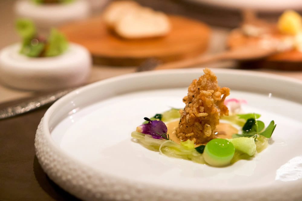 A plated gourmet dish featuring a small portion of fried food atop vegetables and colorful garnishes, presented on a white plate with blurred food in the background.