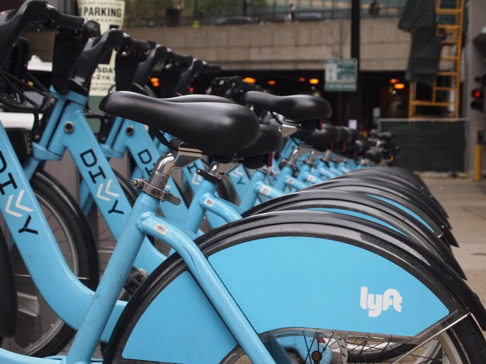 A row of blue Divvy bikes branded with the Lyft logo is lined up at a bike-sharing station on a city sidewalk.