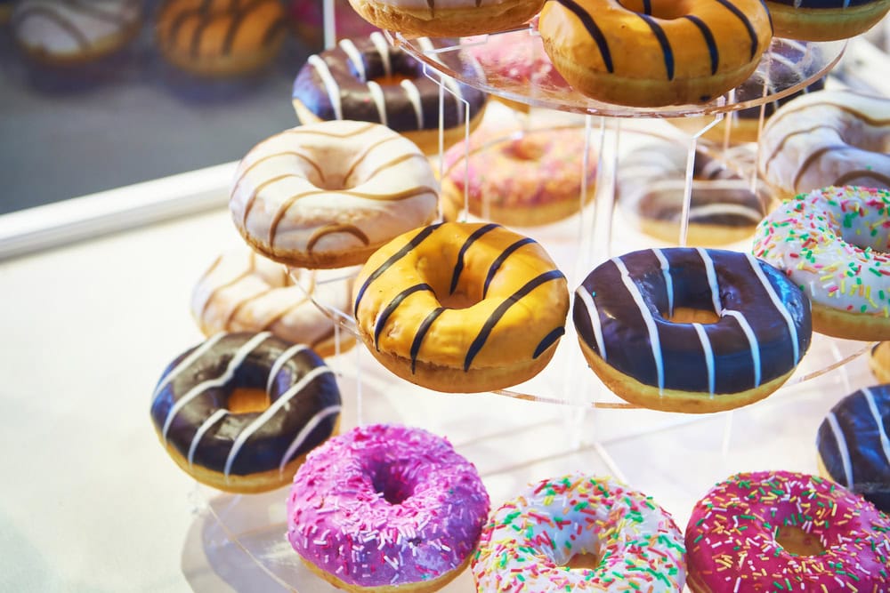 A tiered clear stand displays assorted donuts with colorful icing, glaze, stripes, and sprinkles.