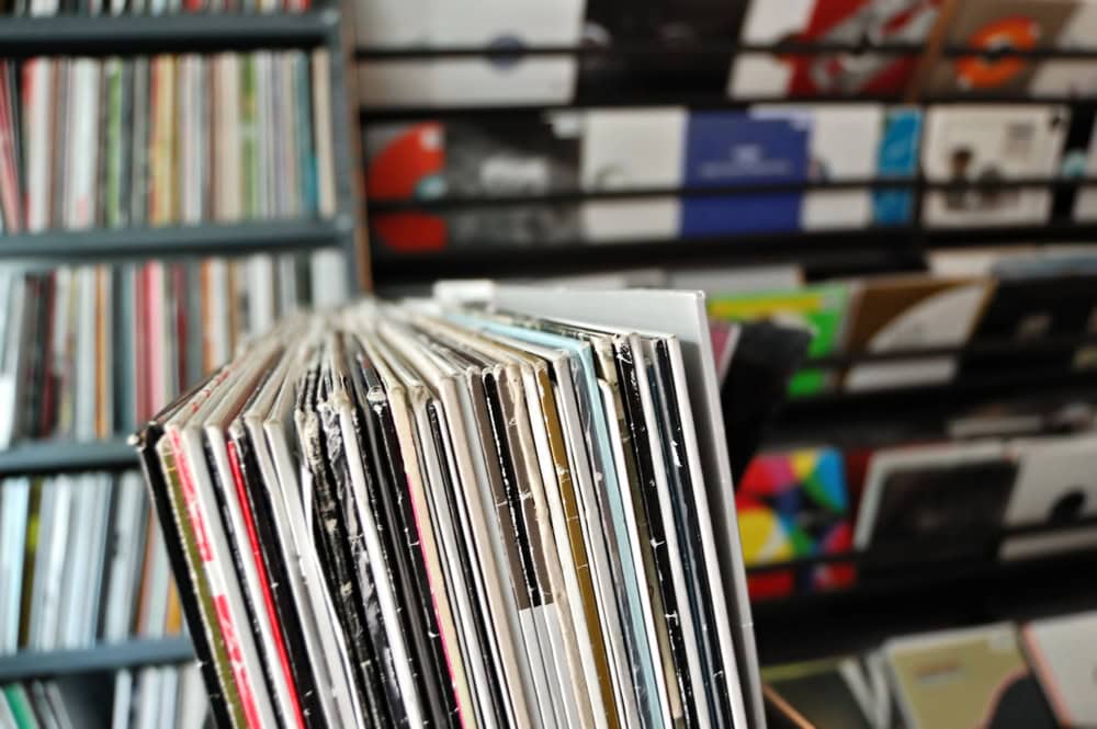A close-up of vinyl records in sleeves displayed upright, with shelves of more records blurred in the background.