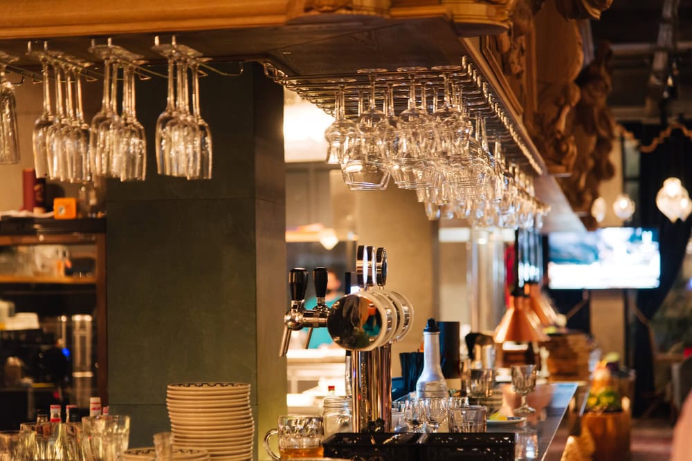 A bar counter with hanging glasses, beer taps, stacked plates, and bottles in a warmly lit restaurant interior.