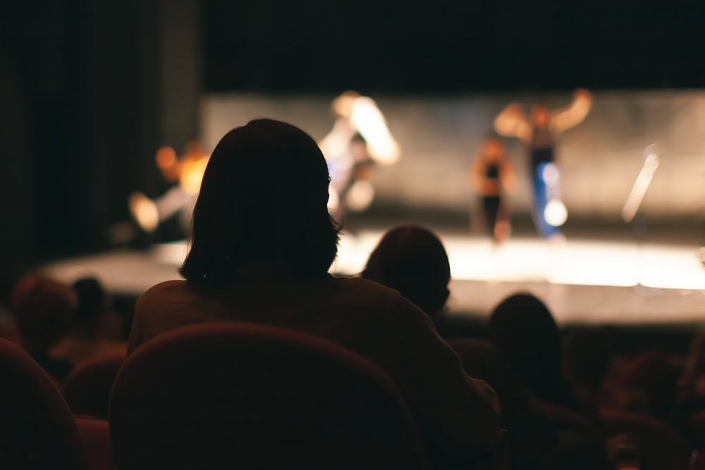 An audience member sits in a dark theater, watching a brightly lit stage where several performers are blurred in motion.