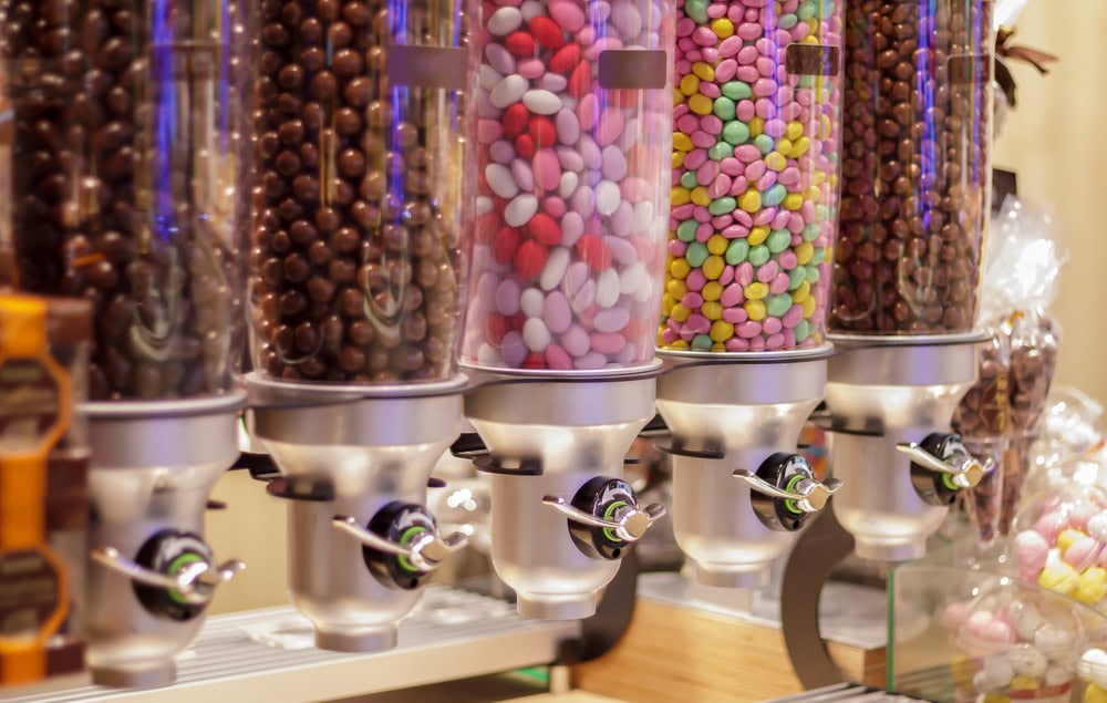 Five clear dispensers filled with assorted colorful candies and chocolate-coated treats are positioned in a row on a counter.