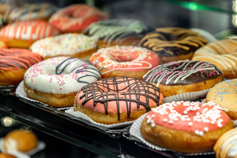 A display case filled with assorted donuts topped with colorful icing, sprinkles, and chocolate drizzles.