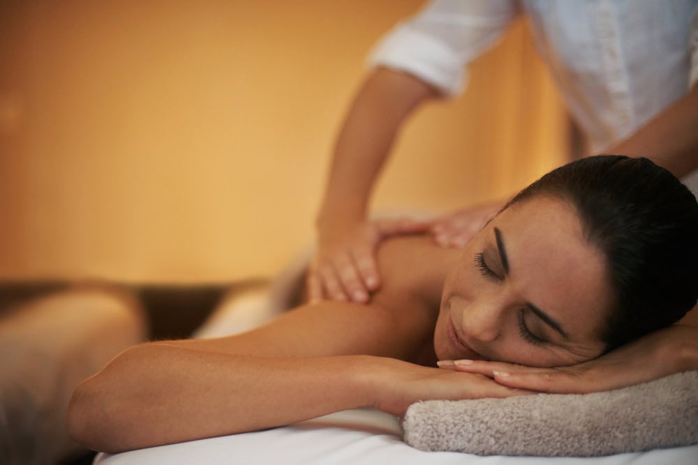 A person lies face down with eyes closed, receiving a back massage from a massage therapist in a softly lit room.