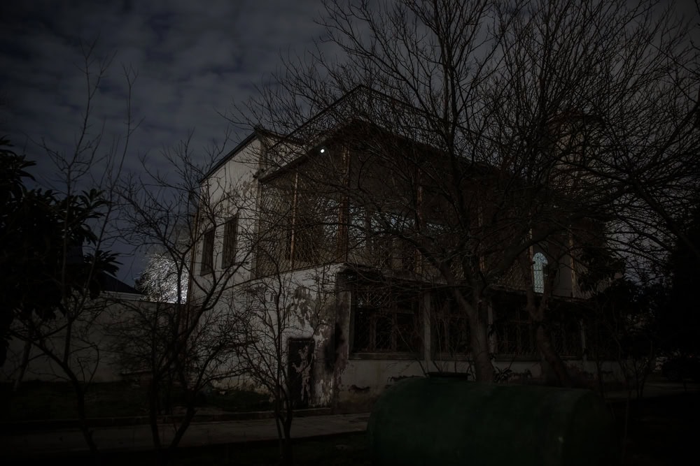 An old, two-story house with peeling paint stands in darkness, surrounded by leafless trees and overgrown grass, with a green tank in the foreground.
