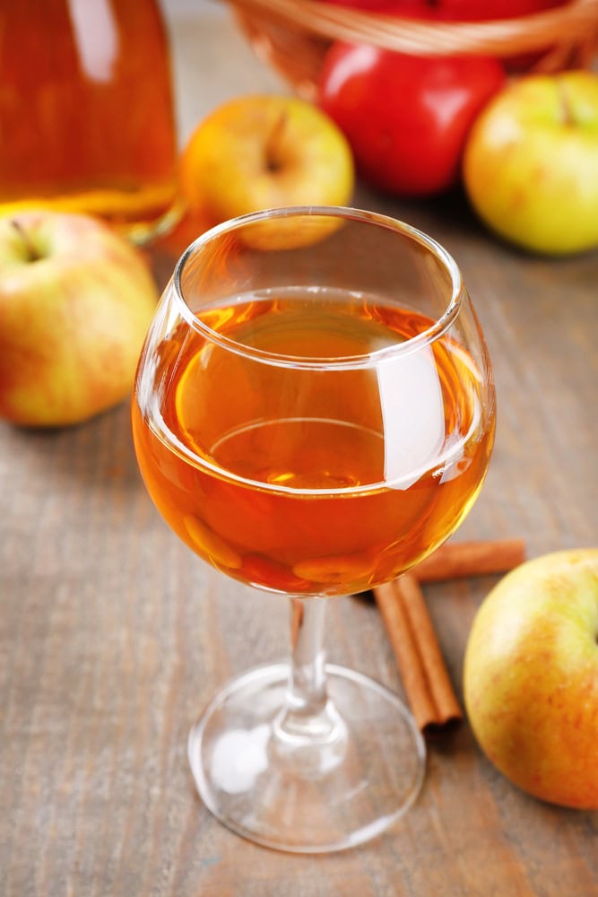 A glass of amber apple cider on a wooden table, surrounded by fresh apples and cinnamon sticks.