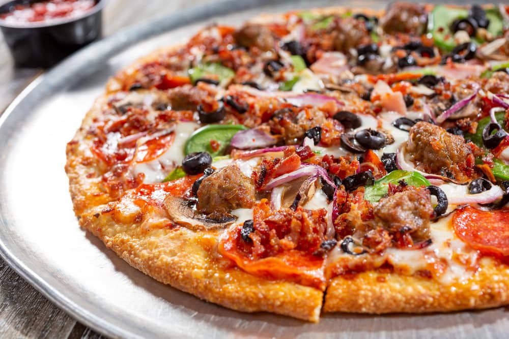 A pizza topped with pepperoni, sausage, bacon, black olives, green peppers, red onions, and mushrooms on a metal tray.