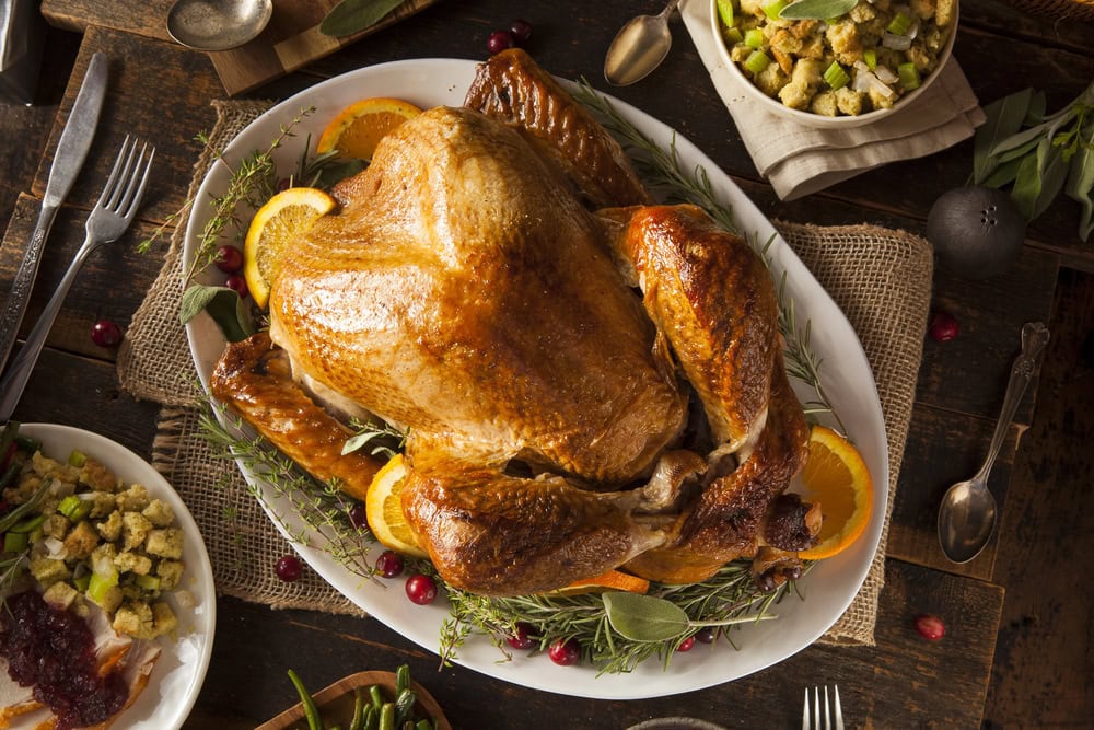 Roast turkey garnished with orange slices and herbs on a white platter, surrounded by bowls of stuffing, cranberry sauce, and green beans on a rustic wooden table.