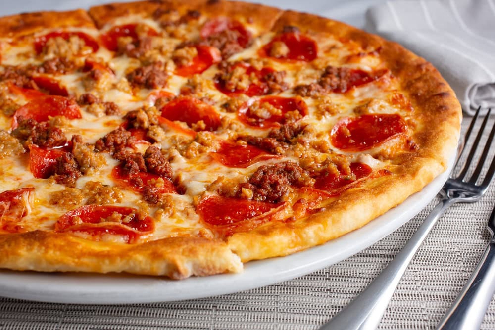 A cooked pizza topped with pepperoni and crumbled sausage sits on a white plate next to a fork and knife on a woven placemat.